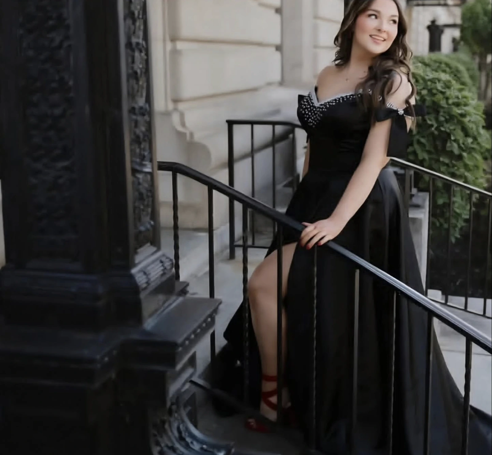 A woman in a black floor-length formal gown with an embellished off-shoulder neckline, bow-like shoulder detail, and high slit stands beside a black railing outside a light stone building.