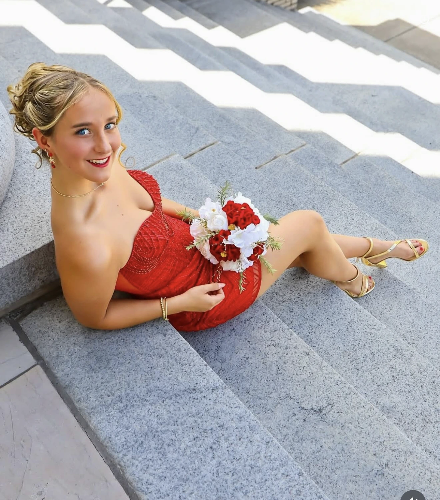 A smiling woman seated on gray stone steps wears a red embellished formal dress, gold strappy heels, and holds a red-and-white bouquet.