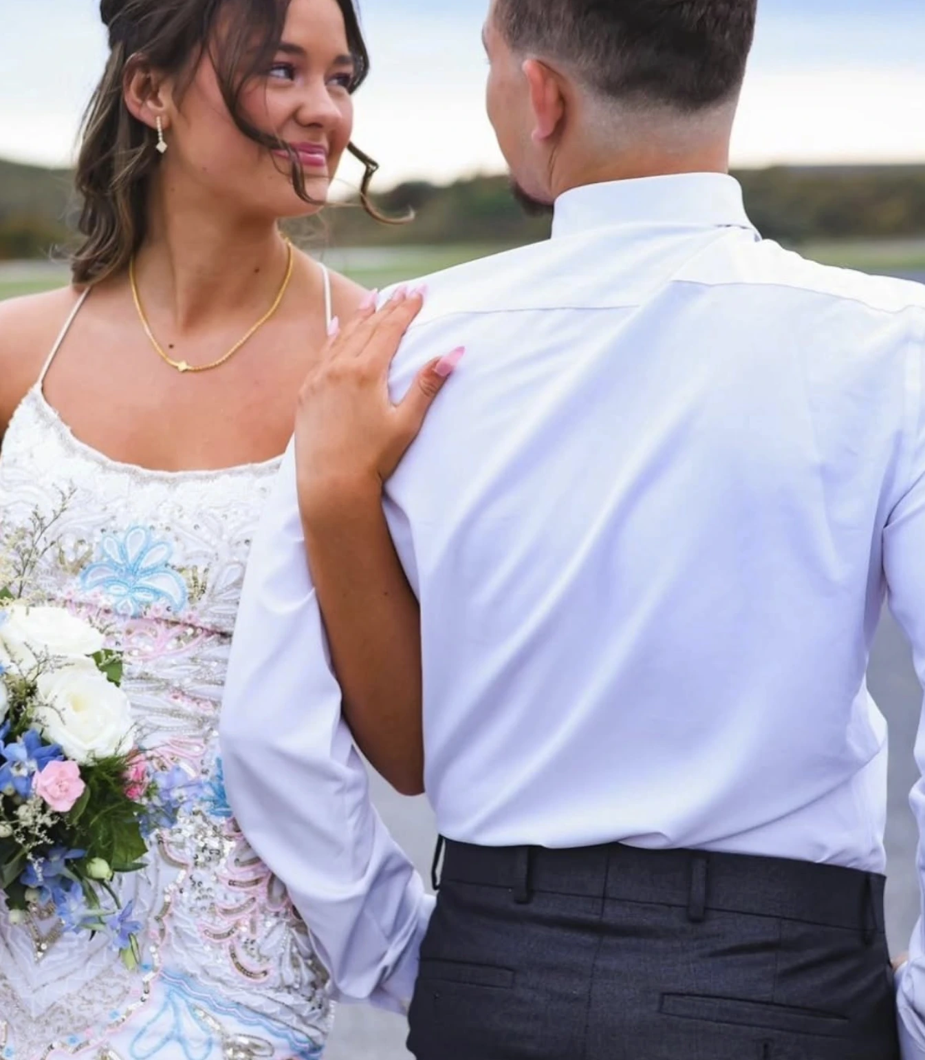 Outdoor formal portrait of a woman in a white spaghetti-strap embellished dress with pastel floral motifs holding a blue, white, and pink bouquet beside a man in a white dress shirt; the woman looks toward him.