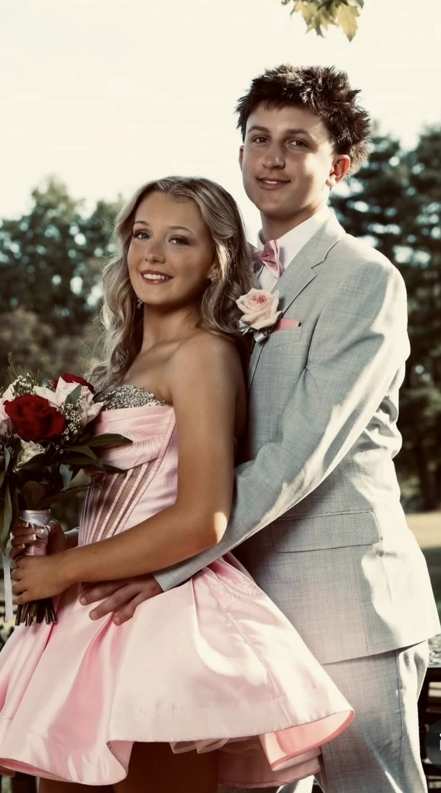A couple poses outdoors, with one person wearing a pale pink strapless short formal dress with a glittering bodice and full flared skirt while holding a bouquet of red and pale flowers; the other wears a light gray suit with a pink bow tie, pocket square, and floral boutonniere.