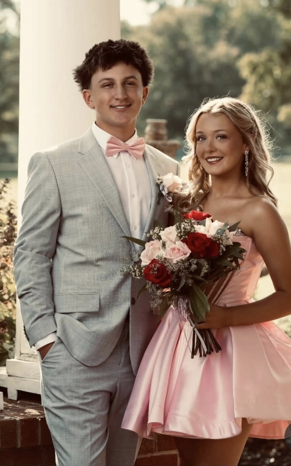 A smiling couple poses outdoors in formalwear: a woman in a pale pink strapless short dress holds a bouquet of red and pale flowers beside a man in a light gray suit with a pink bow tie.