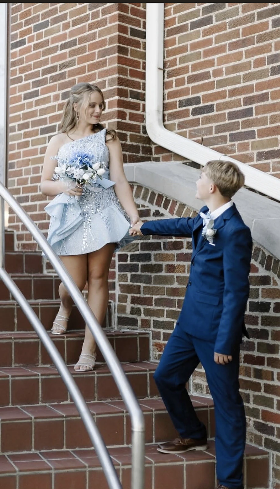 A woman in a light blue embellished short formal dress holds a bouquet on brick steps while a man in a blue suit reaches toward her.