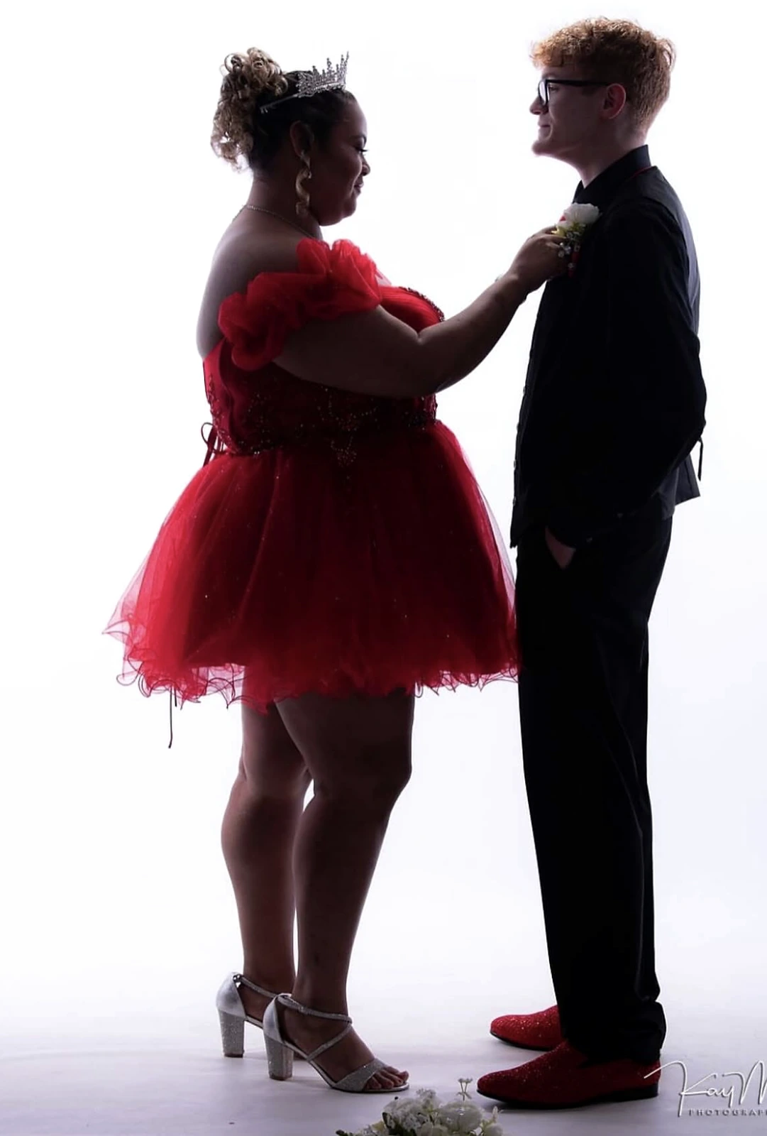 Full-length studio portrait of a woman in a short red tulle formal dress with ruffled off-shoulder sleeves and a tiara standing with a man in a black suit, white boutonniere, and red dress shoes.