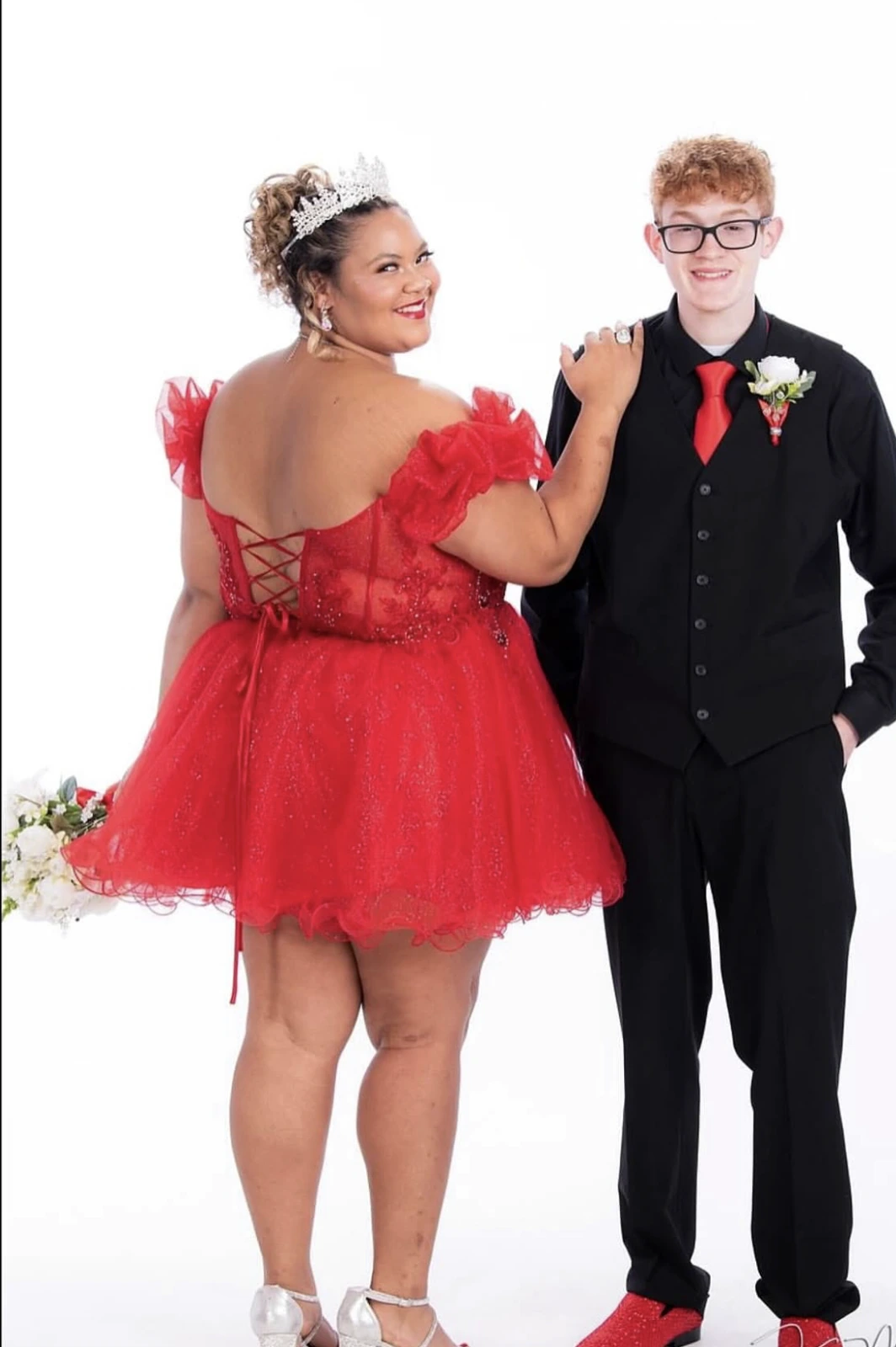 Formal portrait of a smiling woman in a bright red short dress with off-shoulder ruffled sleeves, a lace-up back, sparkling details, and a full tulle skirt, wearing a tiara and silver-toned heels, standing beside a smiling man in black formalwear with a red tie, red shoes, and a boutonniere against a white background.