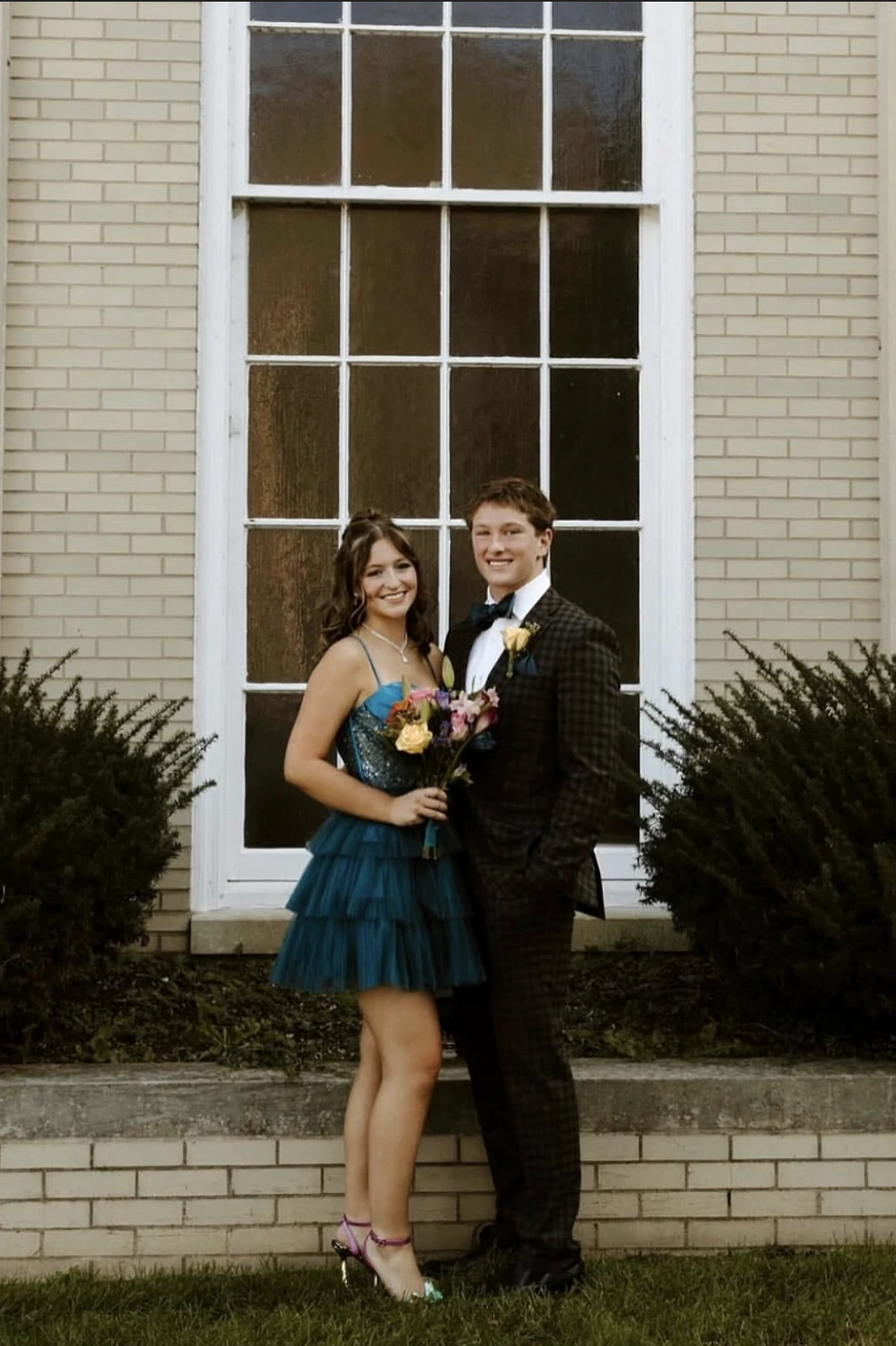 A smiling woman in a teal sparkly spaghetti-strap short dress and purple heels stands beside a smiling man in a dark plaid suit with a bow tie and yellow boutonniere, holding a colorful bouquet in front of a light brick building with a large white-trimmed window.