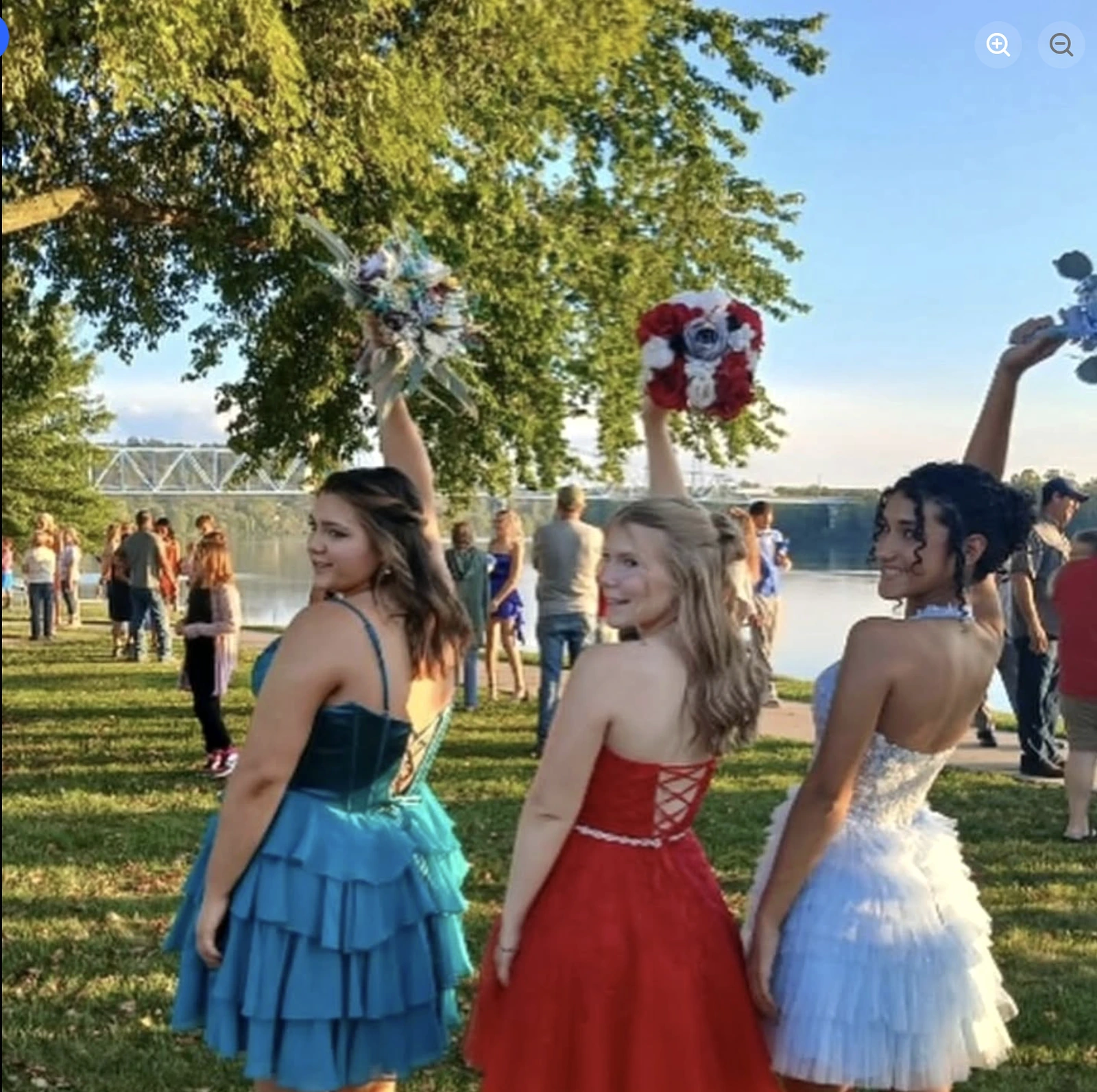 Three women in colorful formal dresses pose on grass beside a river, each holding a bouquet overhead; the left wears a blue-green tiered dress, the center wears a red dress with a lace-up back, and the right wears a pale blue-white tulle dress.