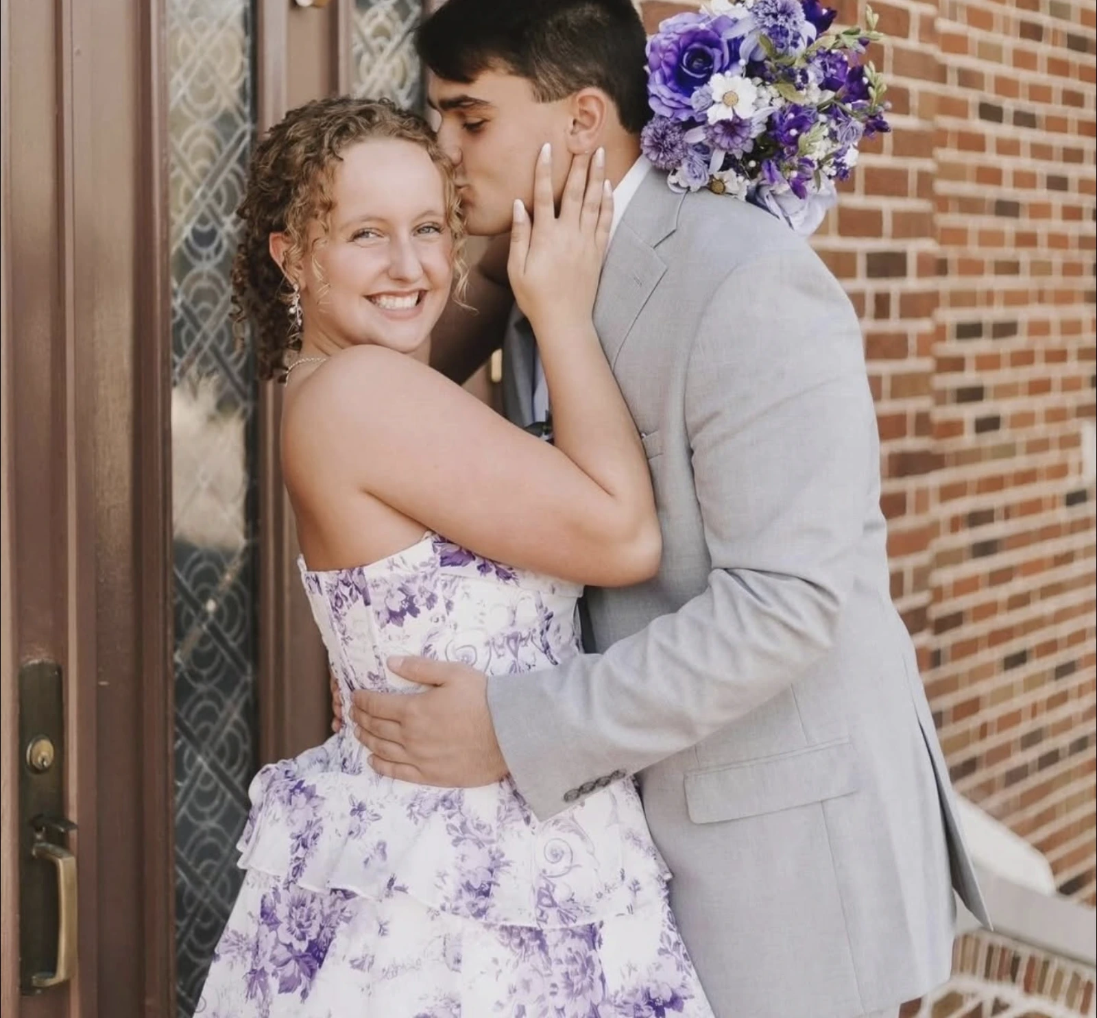 A smiling person in a white-and-lavender floral strapless formal dress poses with a person in a light gray suit at a brick doorway; the suited person kisses the side of their head and holds a purple-and-white bouquet.