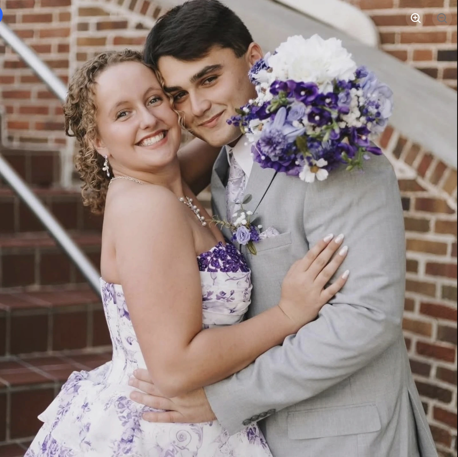 Portrait of a smiling woman in a white-and-lavender floral formal dress embracing a man in a light gray suit, with a purple-and-white bouquet, posed in front of a brick wall.