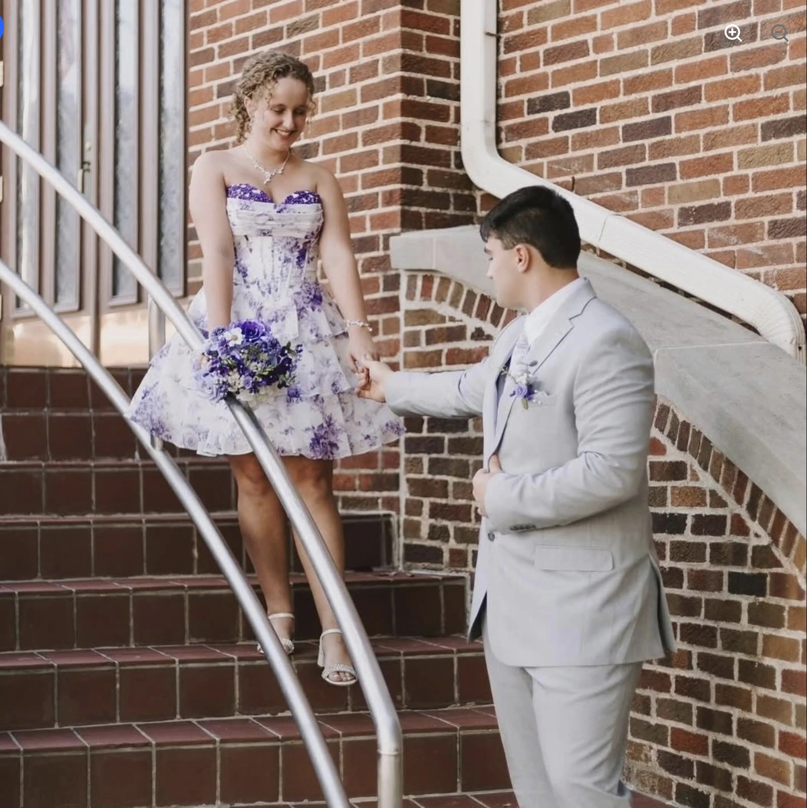 A woman in a strapless white-and-purple floral short formal dress holds a purple-and-white bouquet while standing on brick steps beside a person in a light-colored suit.