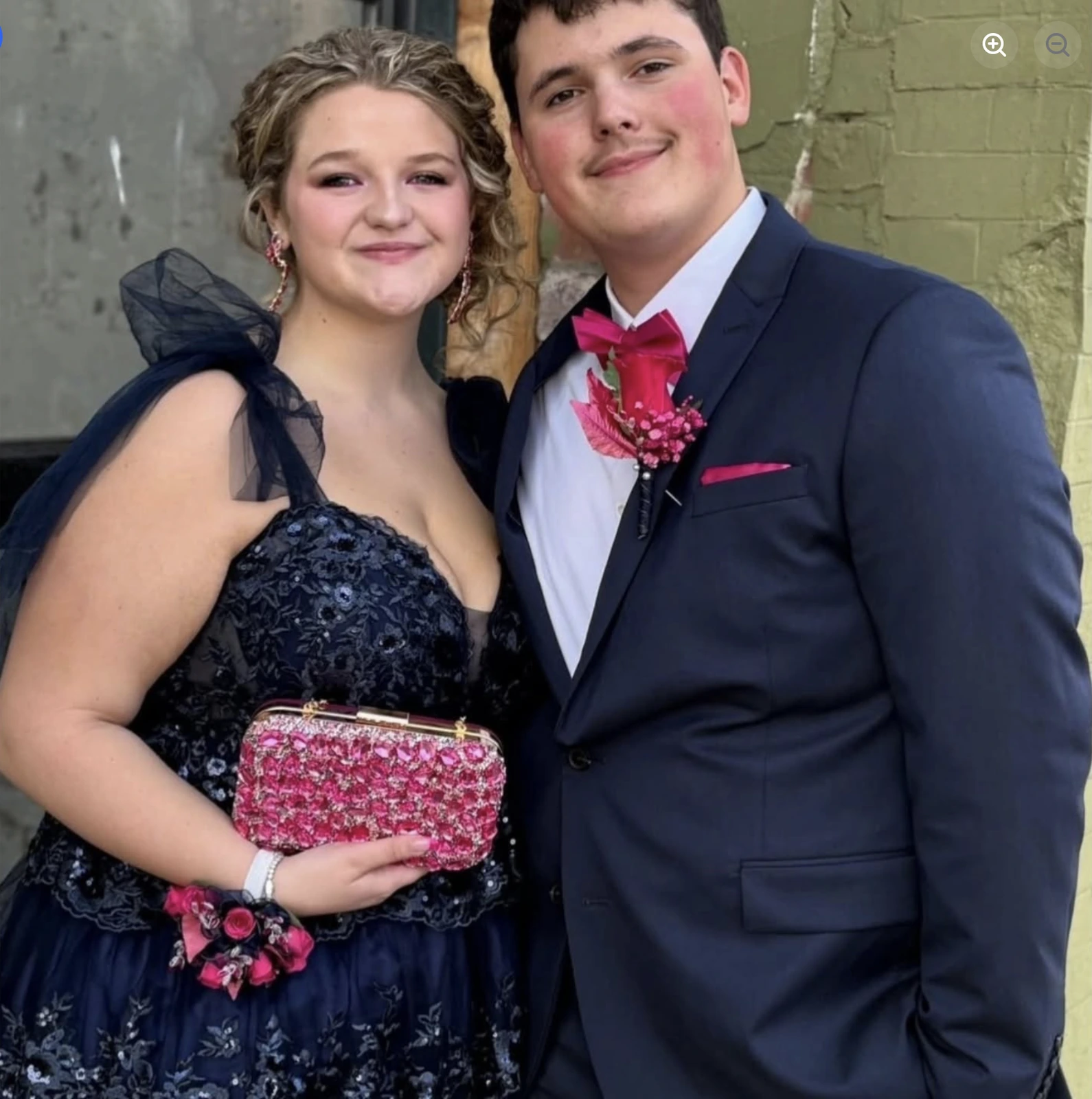 A smiling young woman in a dark navy floral-patterned formal dress with sheer ruffled shoulder details stands beside a smiling young man in a dark navy suit, white shirt, bright pink bow tie, matching pocket square, and boutonniere; she holds a pink patterned clutch.