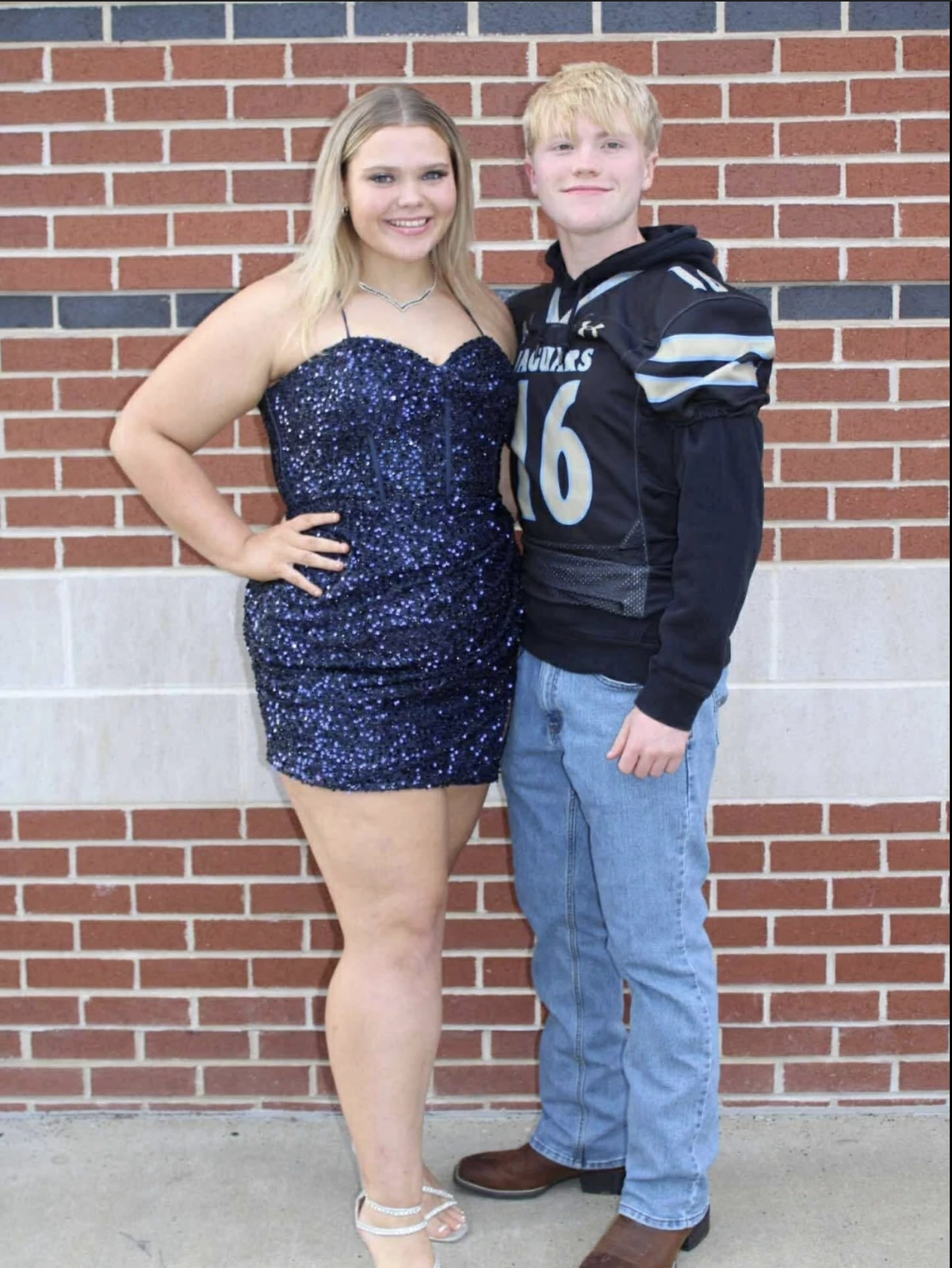 Two people pose in front of a red-and-gray brick wall; a smiling woman wears a short dark navy sequined spaghetti-strap dress and silver sandals beside a person in a black sports jersey and jeans.
