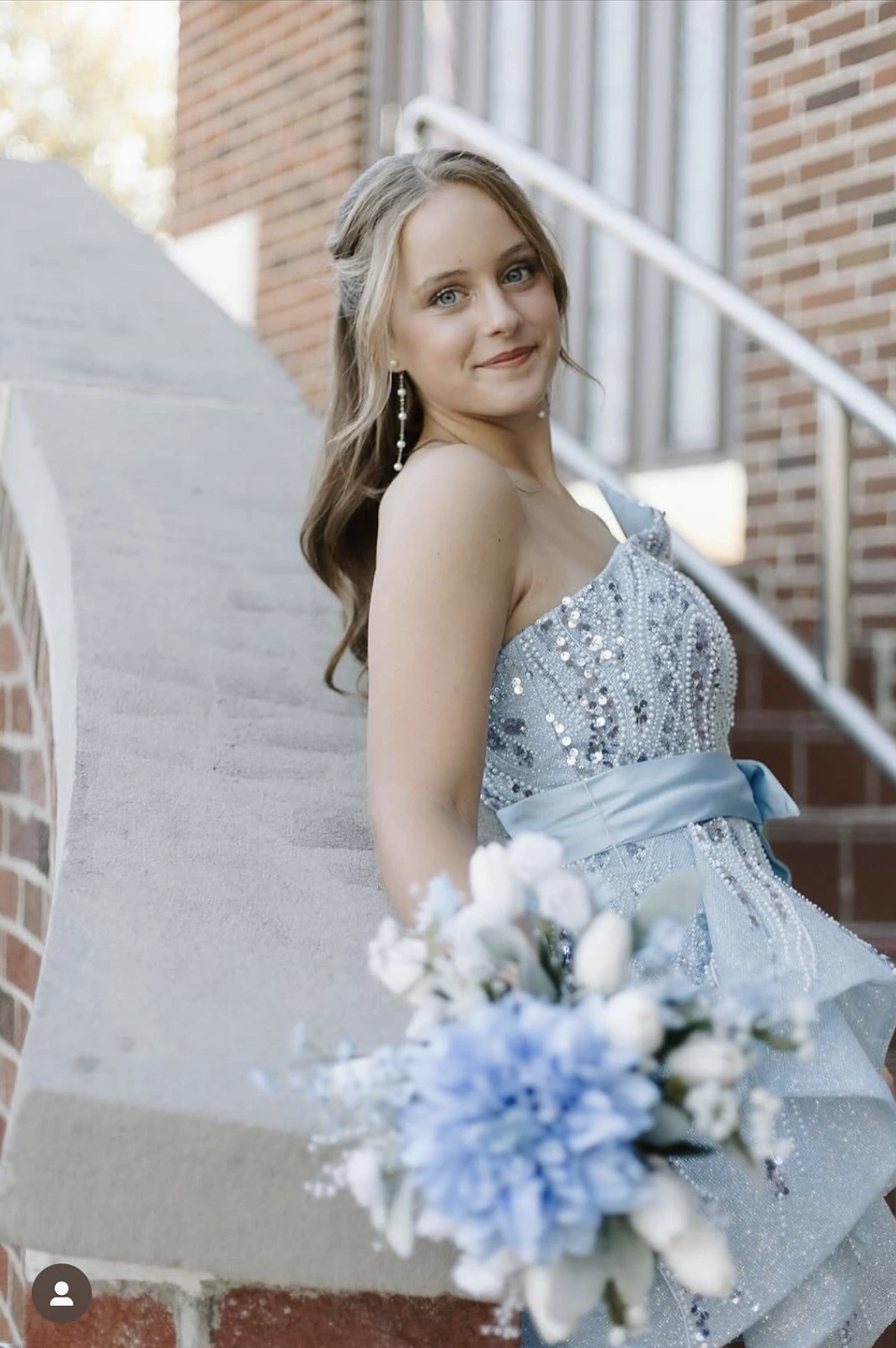 Portrait of a smiling woman with blonde hair wearing a light blue-silver sequined and beaded formal dress with a wide waist sash and layered tulle skirt, seated on outdoor steps beside a brick building; an out-of-focus blue-and-white bouquet obscures the lower foreground.