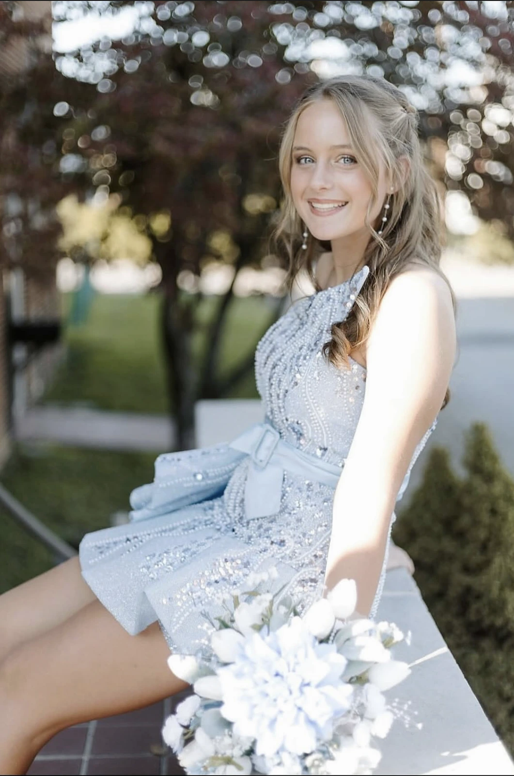 Smiling blonde-haired person seated outdoors, wearing a pale blue sleeveless formal dress with dense sparkling beadwork, sequined detailing, and a large bow at the waist; a light-colored flower bouquet is in the foreground.