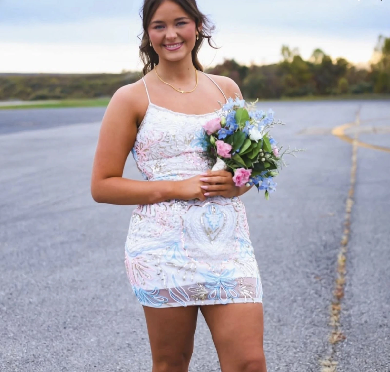 A smiling woman in a pale pastel floral-embellished spaghetti-strap short formal dress holds a pink, blue, and white bouquet outdoors beside a road and body of water.