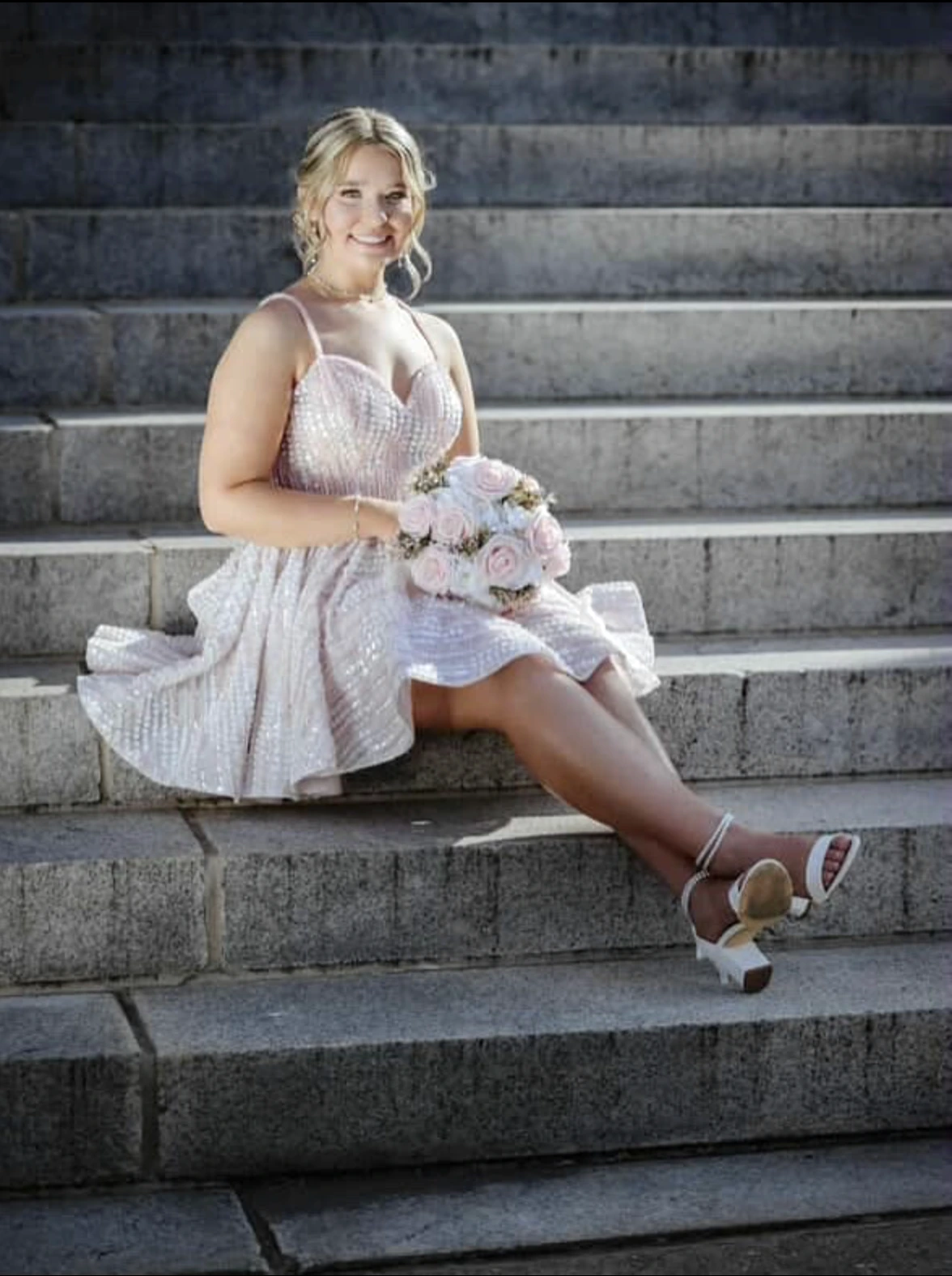 Smiling woman seated on gray stone steps in a pale blush-pink short formal dress with thin straps and a full textured flared skirt, holding a bouquet of pale pink and white flowers and wearing white strappy heeled sandals.