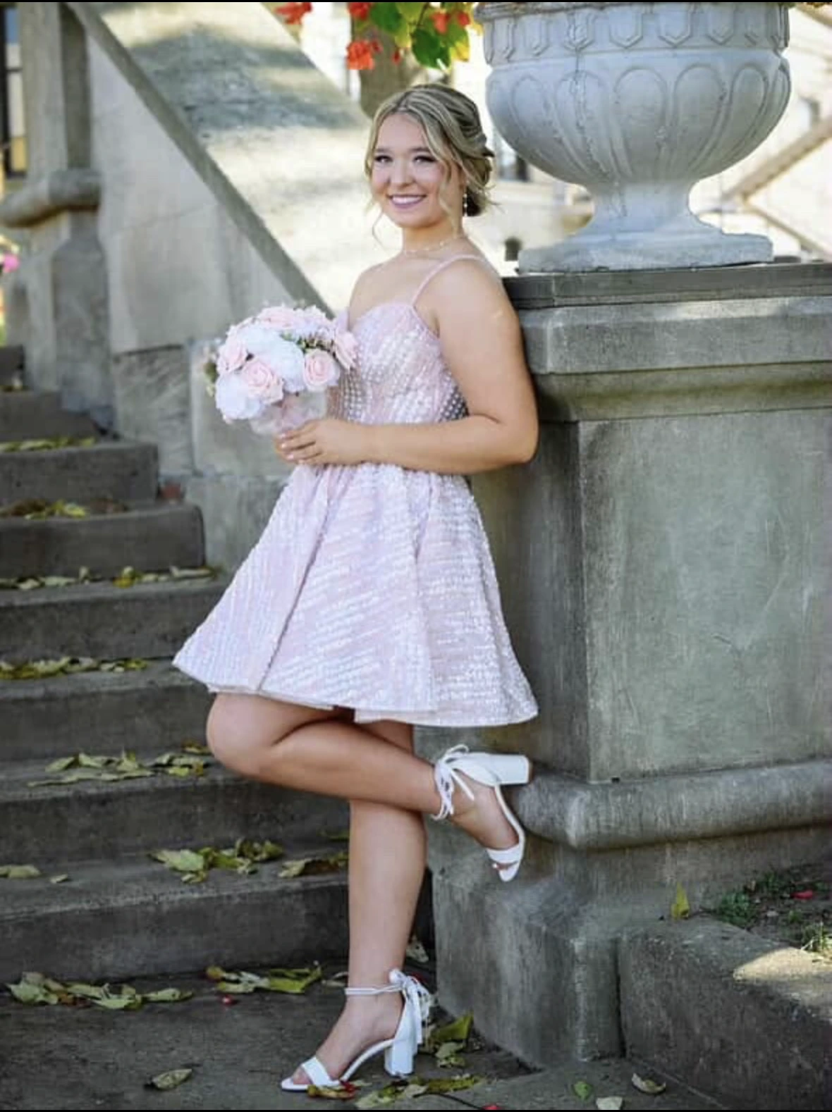 Smiling woman in a light blush-pink short formal dress with thin straps and a textured bodice, holding a pale pink and white bouquet on stone steps beside a large planter; white strappy heels are visible.