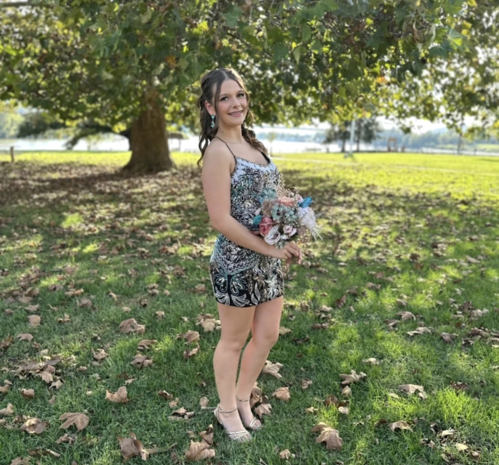 Full-body outdoor portrait of a smiling woman in a sleeveless black-and-white patterned short formal outfit with teal and pink accents, holding a pastel bouquet while standing on grass beneath a leafy tree with water in the background.
