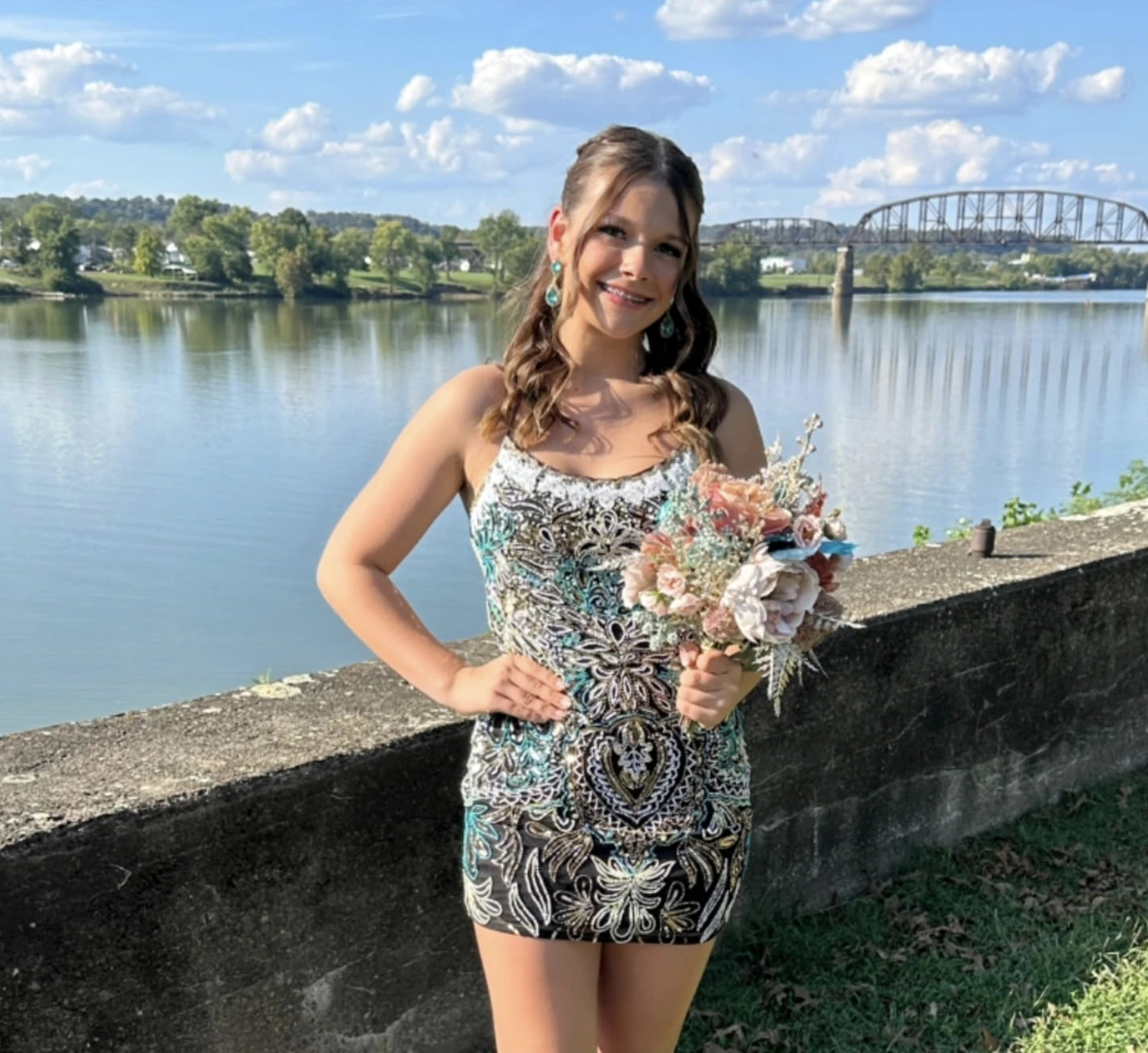Woman with long wavy brown hair wearing a sleeveless black, silver, and teal patterned short formal dress, standing beside a river and holding a pastel bouquet with a steel bridge in the background.