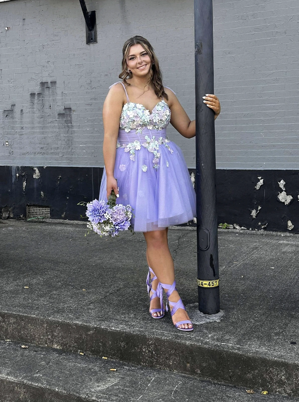 Model wearing a lavender knee-length formal dress with floral appliqué across the bodice and waist and a layered tulle skirt, holding a lavender-and-white bouquet beside a dark utility pole against a gray brick wall.