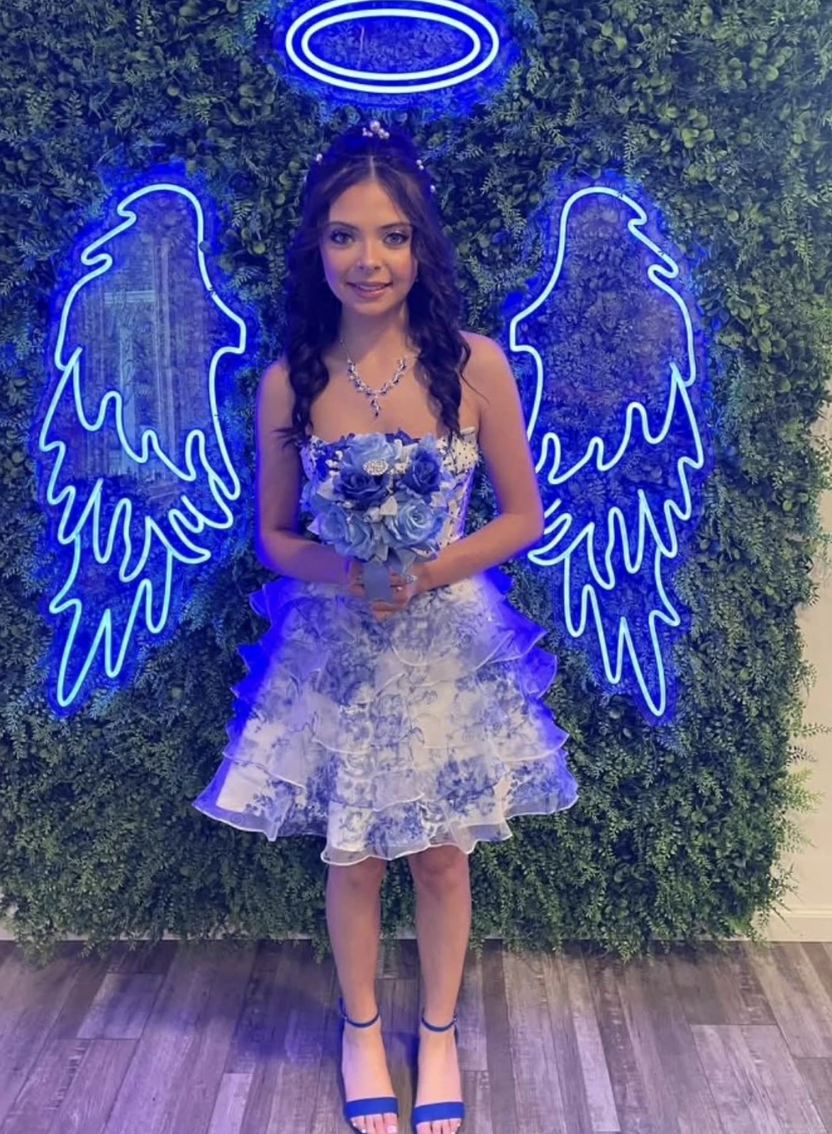 Model facing the camera in a short strapless formal dress with blue-and-white floral detailing on the bodice and a layered ruffled translucent skirt, holding a blue-and-white bouquet in front of a leafy wall with blue neon angel wings; blue ankle-strap heels are visible.