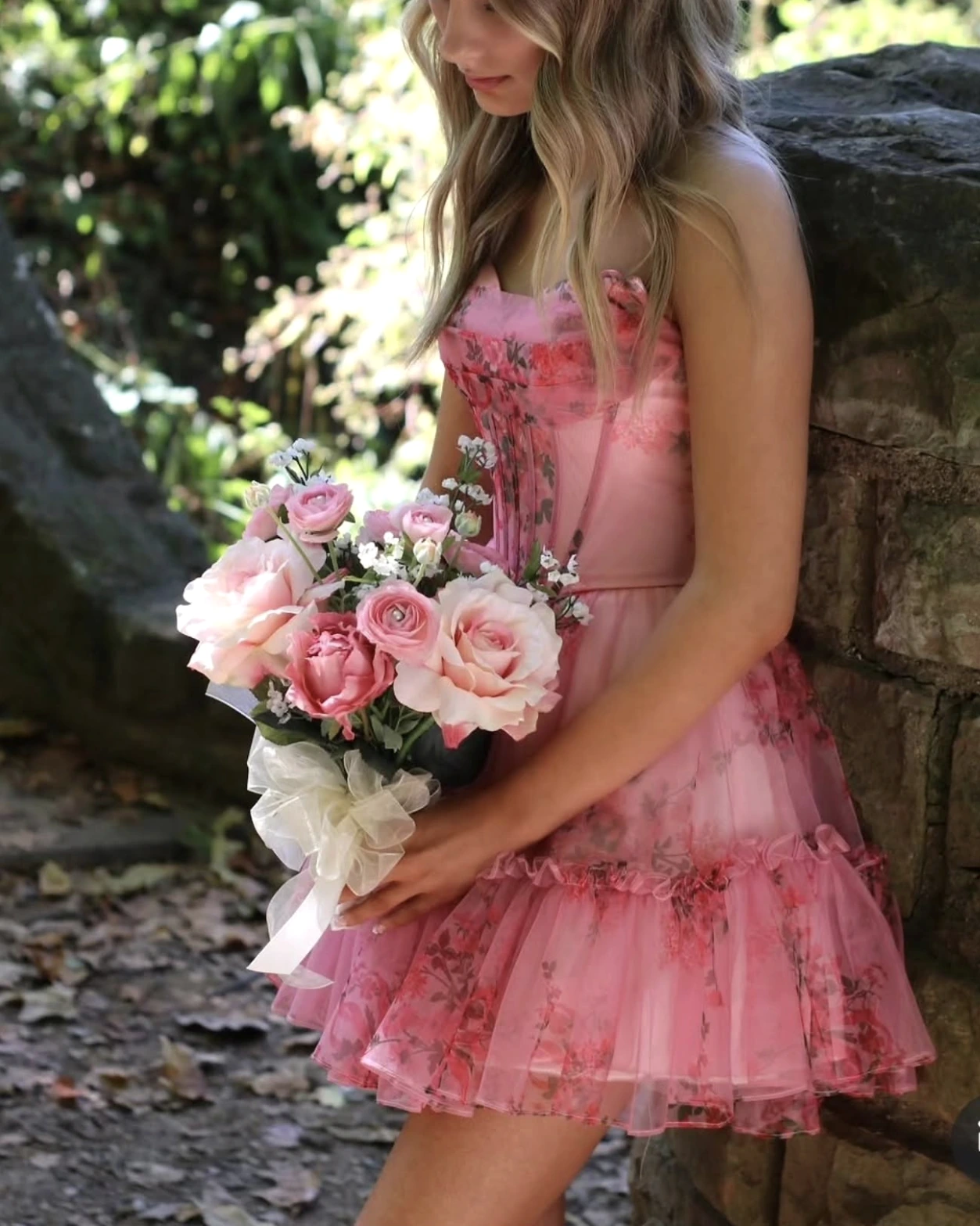 Person with long blonde hair wearing a strapless blush-pink floral formal with a fitted bodice and layered ruffled tulle skirt, holding a pink-and-white bouquet beside a stone wall outdoors; the upper face is partly outside the frame.