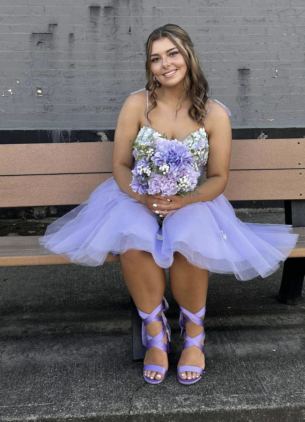 Woman seated on a bench wearing a lavender short formal dress with a floral bodice and full tulle skirt, holding a purple-and-white flower bouquet and wearing matching strappy heels.