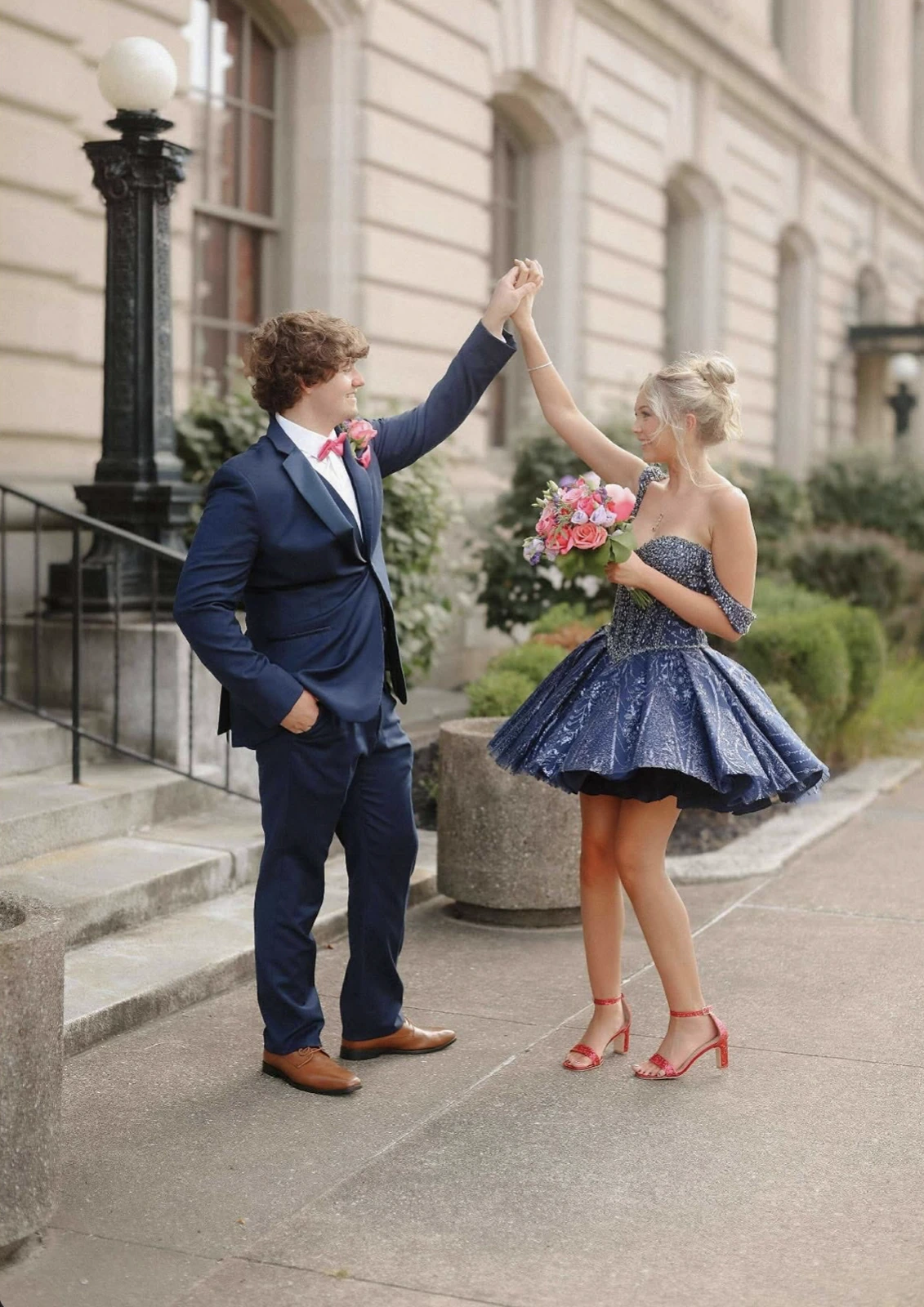Two people in formal attire pose outside a pale stone building, holding hands overhead; one wears a navy suit with a pink bow tie and boutonniere, and the other wears a short blue embellished dress with a full skirt, red heeled sandals, and holds a colorful bouquet.