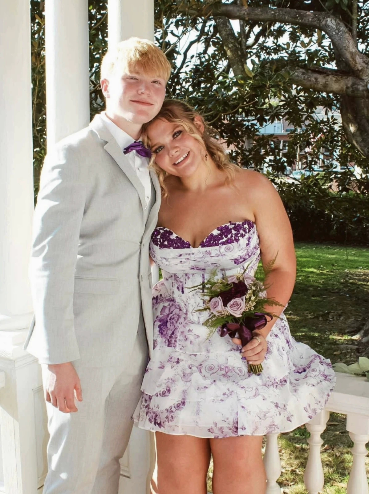 Couple posing outdoors beside white columns and leafy trees; woman in a strapless white short dress with lavender and purple floral print holding a bouquet, man in a light suit with a white shirt and purple bow tie.