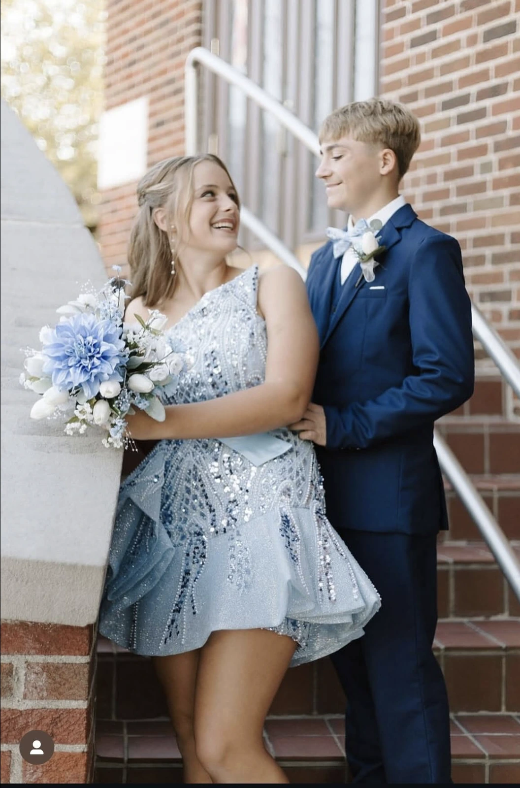 A smiling couple poses on brick steps outdoors; the woman wears an icy blue and silver embellished sleeveless short formal dress and holds a pale blue and white bouquet, while the man wears a navy suit with a light blue bow tie and boutonniere.