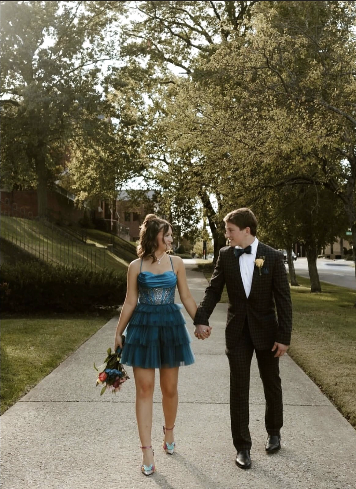 Two young people in formalwear walk hand-in-hand on a tree-lined sidewalk: a woman wears a teal short dress with a tiered tulle skirt and carries a bouquet, while a man wears a dark patterned tuxedo with a bow tie and yellow boutonniere.
