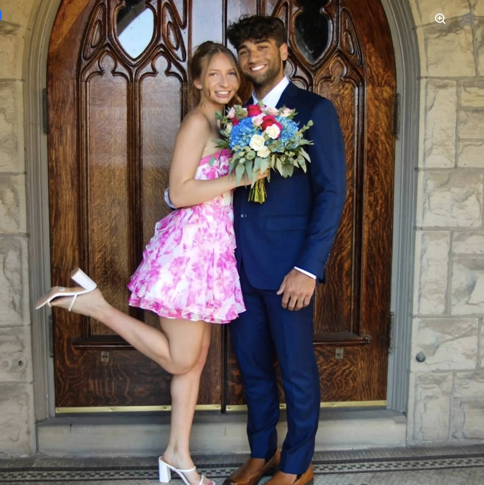 A woman in a pink-and-white floral short formal dress and white heels stands beside a smiling man in a navy suit holding a colorful bouquet in front of carved wooden doors.