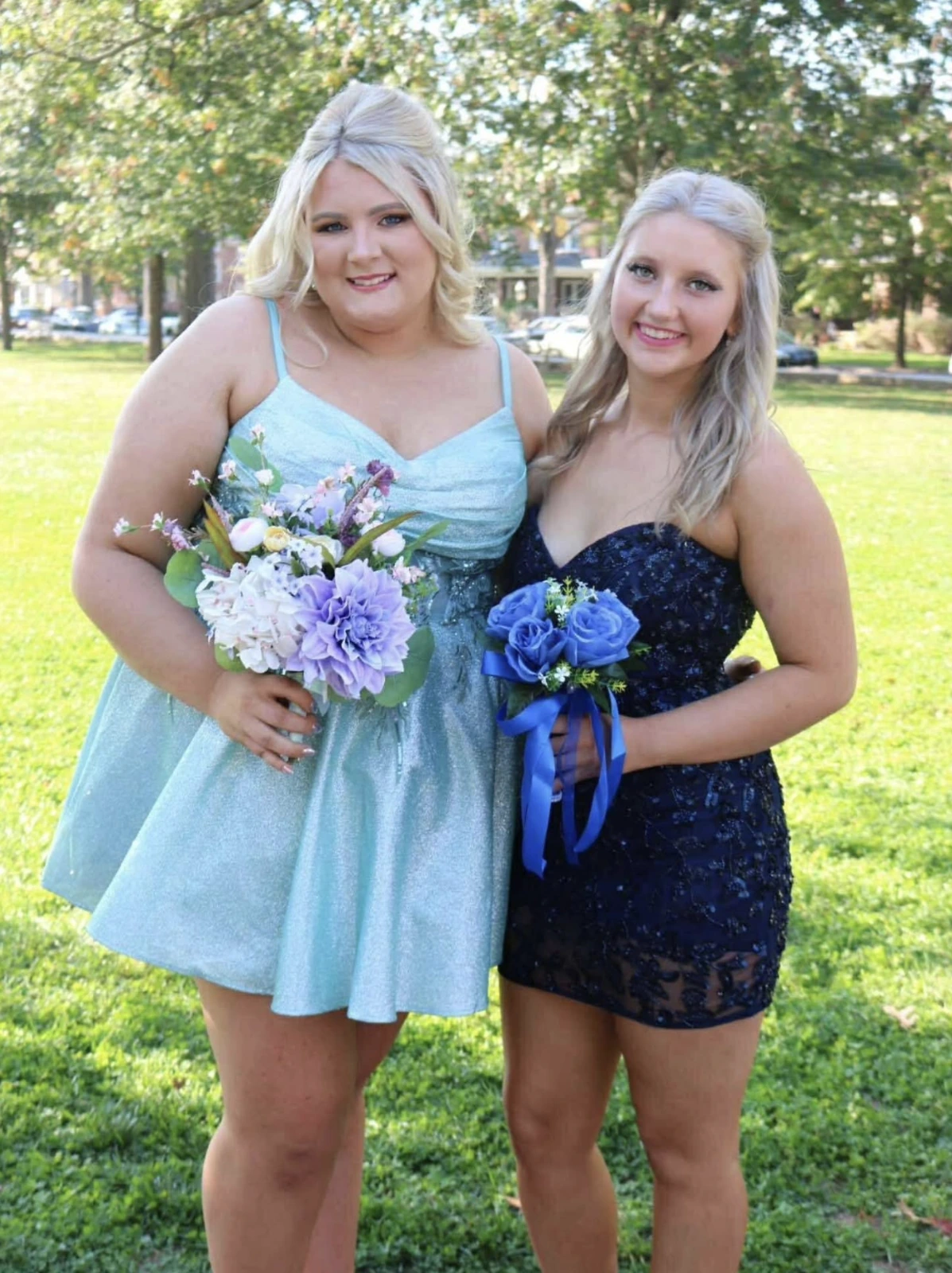 Two smiling women stand together on a grassy lawn, one in a light blue short dress and one in a navy short dress, each holding a floral bouquet, with leafy trees and parked cars in the background.