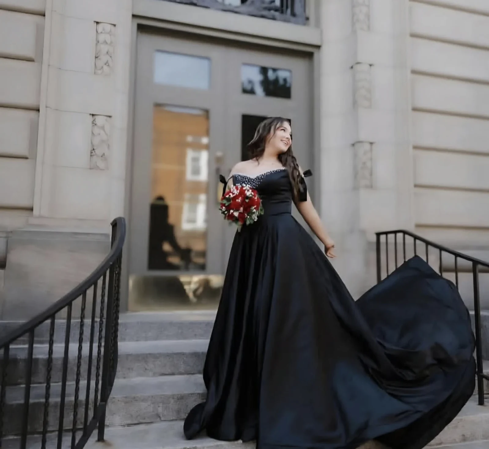 Woman on outdoor steps wearing a black off-the-shoulder floor-length gown with a full skirt and long train, holding a red-and-white bouquet.
