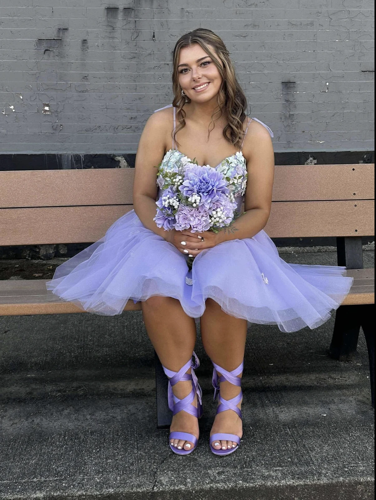 Seated person in a short lavender tulle dress with a floral-patterned bodice, holding a purple-and-white bouquet and wearing purple strappy heels.
