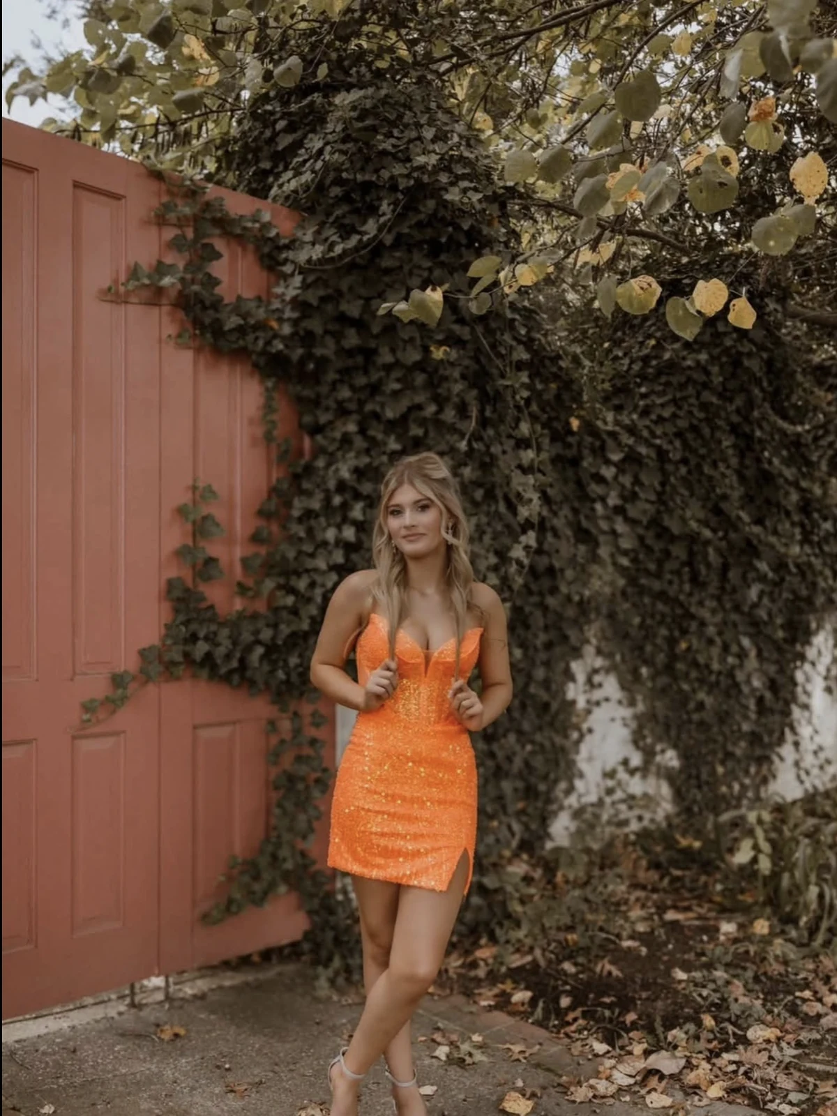 Person in a bright orange sparkly short dress with thin straps and a structured bodice in an outdoor garden setting.