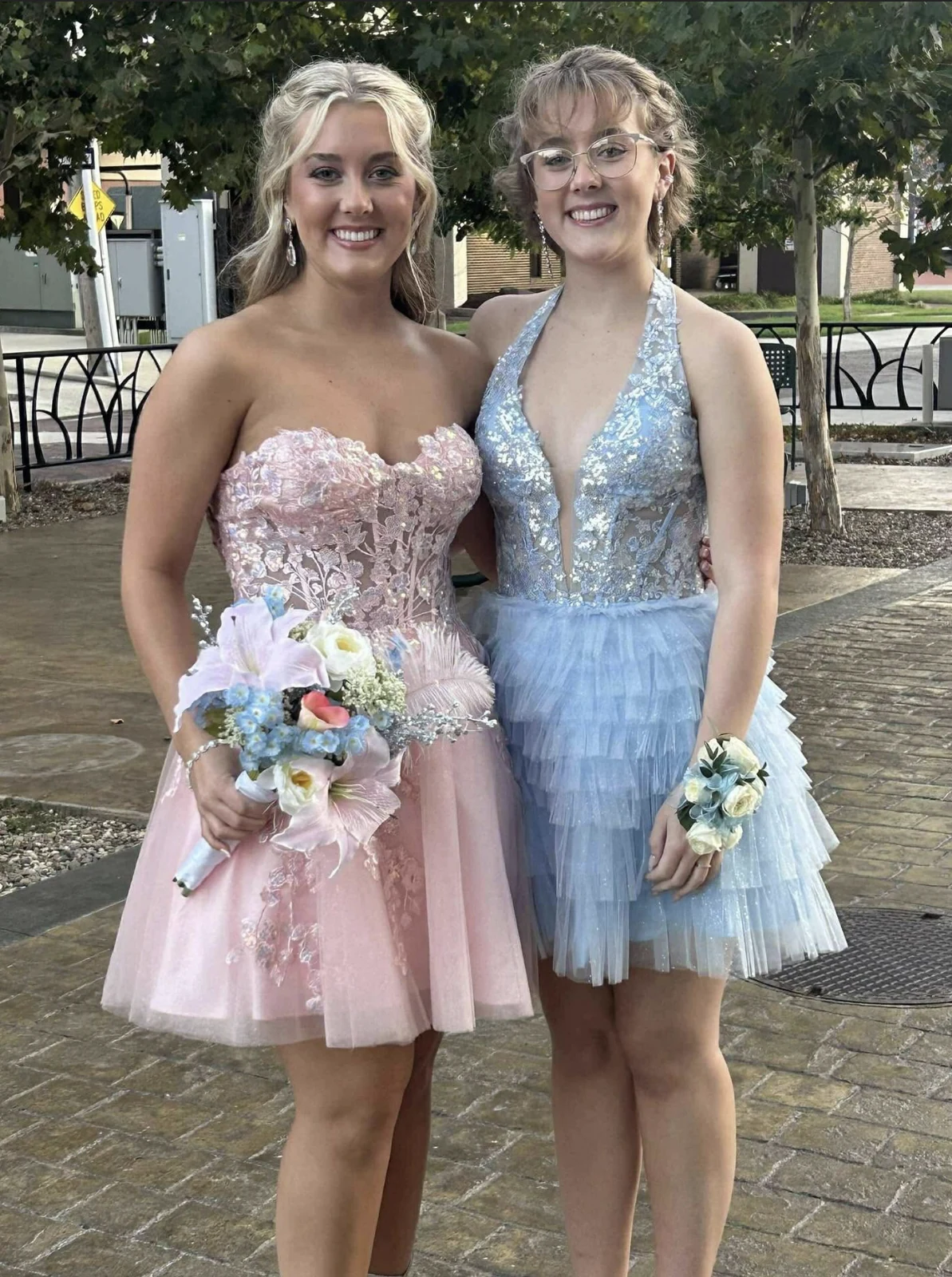 Two people outdoors in a pale pink strapless floral-appliqué short dress and a light blue halter short dress with layered tulle detailing.