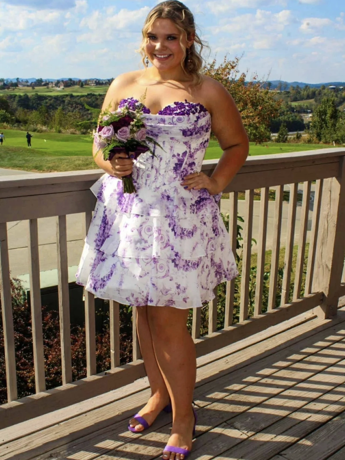 Person outdoors in a short strapless white dress with a purple floral print and flared skirt, holding a bouquet and wearing purple open-toe heels.